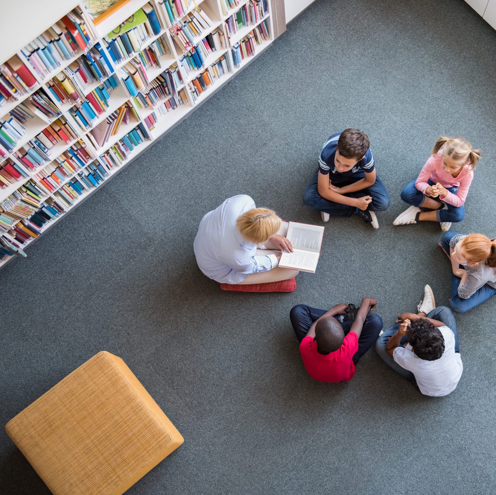 Eine Gruppe von Kinder neben einem Bücherregal. Eine Person liest vor. Ein gelber Hocker in der linken Bildecke.