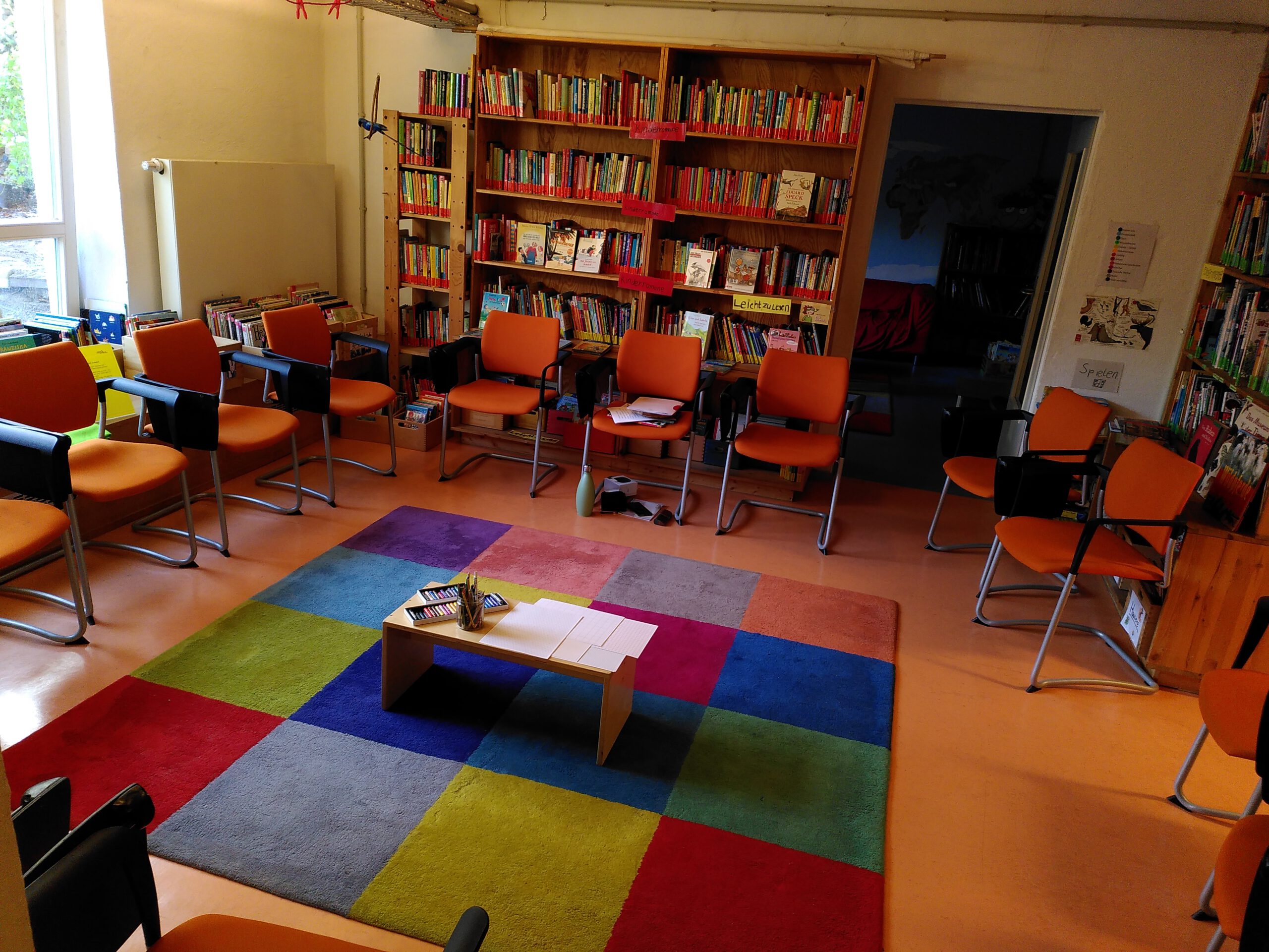 Regale mit Büchern im Hintergrund, im Vordergrund ist ein kleiner Tisch mit papieren und Stiften, drum herum ein Kreis mit leeren Stühlen