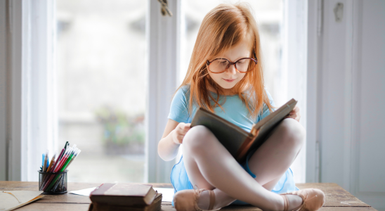 Kind mit Brille sitzt im Schneidersitz vor einem Fenster und liest in einem Buch.