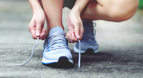 Man sieht die Füße einer Person. An den Füßen sind Sportschuhe. Die Person hockt auf einer Straße und bindet sich gerade einen der Schuhe zu.