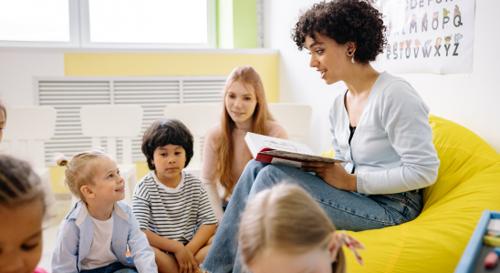 Eine Frau sitzt auf einem gelben Sitzsack und hält ein geöffnetes Buch in der Hand. Vor ihr auf dem Boden sitzen Kinder.