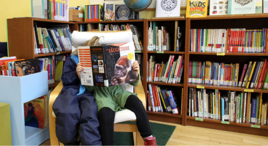 In der Schulbibliothek der Nürtingen Grundschule lesen zwei Kinder, die zusammen auf einem Stuhl sitzen. Im Hintergrund sind ein gefülltes Bücherregal und ein Podest zu sehen.