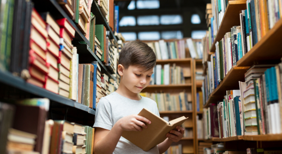 Junge mit offenen Buch in den Händen zwischen Bücherregalen