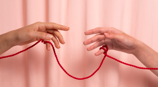 Zwei Hände sind mit einem roten Faden miteinander verbunden. Im Hintergrund ist ein rosa Vorhang zu sehen.