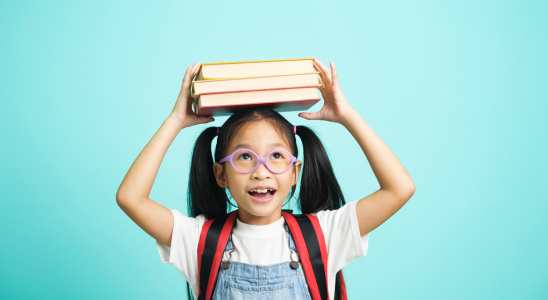 Kind hält drei Bücher mit beiden Händen über den eigenen Kopf. Das Kind trägt einen Rucksack und steht vor einem türkisfarbenen Hintergrund.