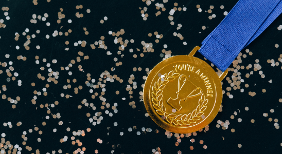 schwarzer HIntergund mit silbernem und goldenen Konfetti unscharf im Hintzergrund. Im Vordergrund auf der rechten Seite befindet sich eine goldene Medaille mit der Inschrift "You're a winner". An der Medaille befindet sich ein blaues Band, was am rechten/oberen Bildrand abgeschnitten wird.