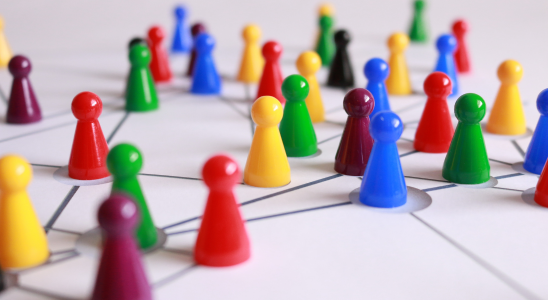 Mehrere bunte Spielfiguren stehen auf Kreisen, die auf einem weißen Blatt angeordnet sind. Die Kreise sind mit Linien verbunden. Die Figuren sind in verschiedenen Farben wie Rot, Gelb, Blau und Grün angeordnet und symbolisieren ein Netzwerk oder eine Vernetzung von Personen.