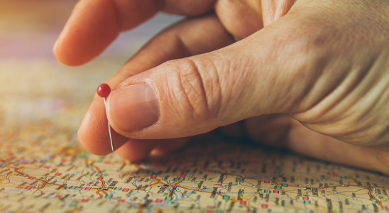 Rechte Hand liegt auf einer Landkarte. Eine Pin-Nadel mit rotem Kopf wird von Daumen und Mittelfinger über der Karte gehalten. Von der Hand ist nur ein kleiner Teil sichtbar, der Rest ist am oberen und rechten Rand des Bildes angeschnitten.
