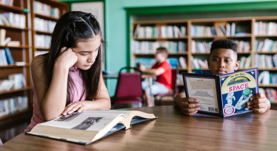 An einem Holztisch in einer Bibliothek sitzen zwei Kinder und lesen. Das Mädchen auf der linken Seite blättert in einem großen, aufgeschlagenen Buch und stützt den Kopf auf die Hand. Der Junge rechts hält ein buntes Kinderbuch mit dem Titel „Space“ in den Händen und liest darin. Im Hintergrund sind hohe Bücherregale voller Bücher zu sehen, außerdem sitzt ein weiteres Kind an einem Tisch und liest. Die Atmosphäre wirkt ruhig und konzentriert, typisch für eine Bibliothek.