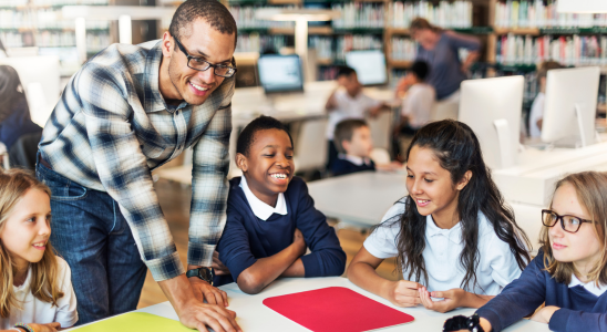 Vier Kinder sitzen um einen Tisch herum. Eine erwachsene Person beugt sich über den Tisch. Im Hintergrund sind unscharf weitere Tische mit Menschen daran zu sehen.