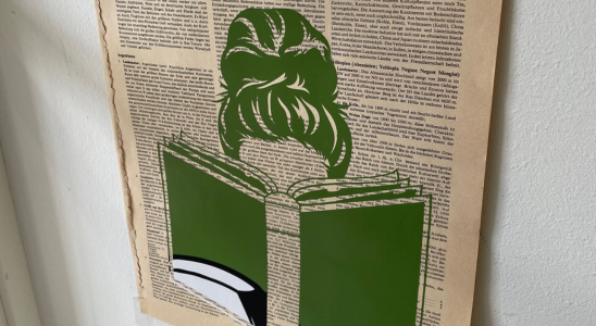 Eine alte Buchseite hängt an einer Wand. Auf der Buchseite wurde mit grüner Flexfolie eine Frau mit einem aufgeschlagenem Buch geplottet.