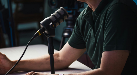 männlicher Oberkörper an einem Tisch sitzend; auf dem Tisch steht ein Mikrofon, welches zur Person ausgerichtet ist; Hintergrund ist sehr dunkel