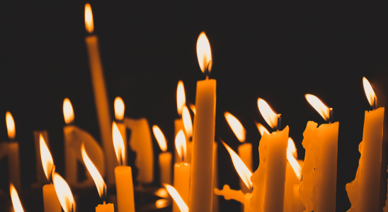 Schwarzer Hintergrund. Davor stehen knapp 20 Stabkerzen, die alle brennen und unterschiedlich weit heruntergebrannt sind.