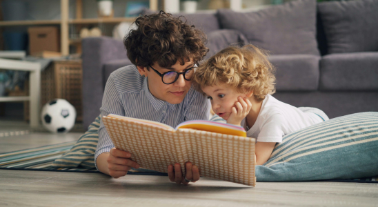 Eine Person liegt auf dem Bauch auf dem Boden und liest einem Kind, was auch auf dem Bauch liegt aus einem Buch vor. Beide liegen auf einem Teppich vor einer Couch. Hinter der Couch stehen ein Tisch und ein Regal. Spielzeug ist unscharf im Hintergrund zu sehen.