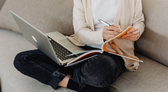 Frau sitzt auf einer hellen Couch. Si ehat einen Laptop auf dem rechten Knie. Auf dem linken Knie liegt ein Heft, in das sie mit einem Kuli reinschreibt.