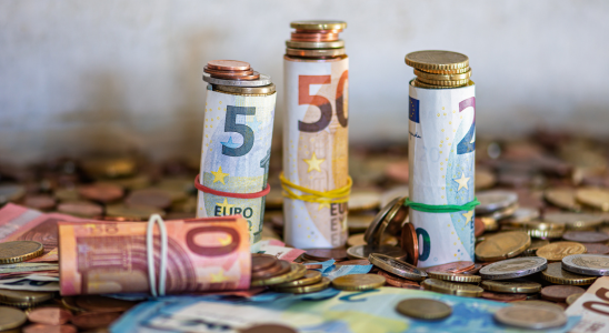 Drei zusammengerollte Geldscheine stehen auf einem Tisch, eine Rolle liegt auf dem Tisch im Vordergrund. Um die Geldscheine herum liegen viele Münzen. Einige Münzen sind auch auf die stehenden Geldrollen platziert.