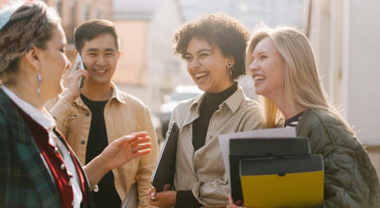 vier Menschen stehen draußen und unterhalten sich angeregt. Im Hintergrund kann man Sonne erahnen. Eine Person hat einen Bloh oder Buch in der Hand. Alle Personen sieht man nur bis zur Brust.