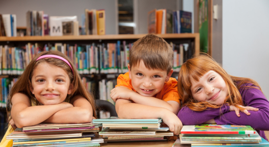 Drei Kinder lehnen mit Armen und Köpfen auf Bücherstapeln. Im Hintergrund sind verschwommen einige Buchregale zu erkennen.