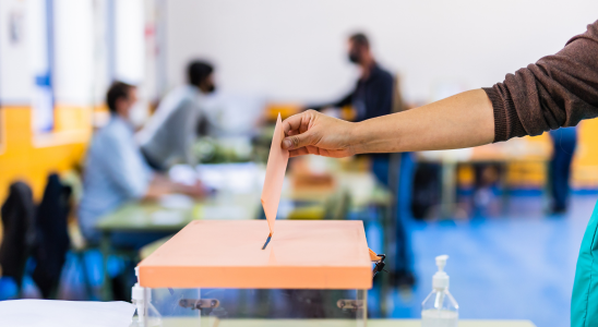 Ein Zettel wird in eine Wahlurne gesteckt. ZU sehen ist der Unterarm der wählenden Person und die obere Seite der Urne. Im Hintergrund sind sehr unscharf drei Personen zu sehen.