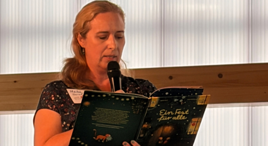 Maike Harel steht vor einer halbtransparenten Wand und liest aus ihrem Buch "Ein Fest für alle".