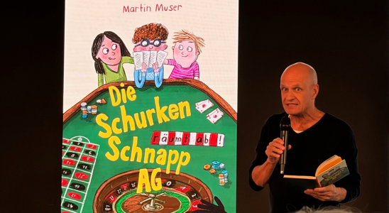 Martin Muser steht vor einem großen Bildschirm. Darauf ist das Cover seines Buches "Die Schurken Schnapp AG" zu sehen.