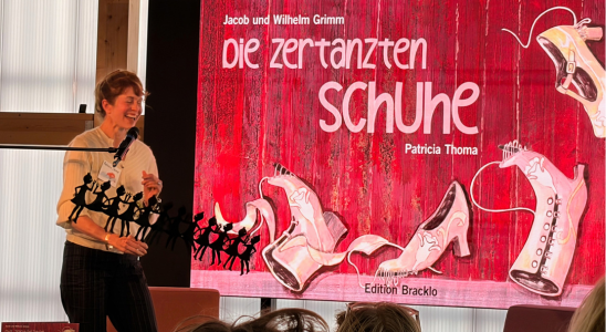 Patricia Thoma steht vor einem großen Bildschirm. Darauf ist das Cover ihres Buches "Die zertanzten Schuhe" zu sehen.