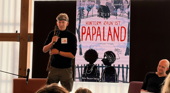 Ulrich Fasshauser steht vor einem großen Bildschirm. Darauf ist das Cover seines Buches "Hinterm Zaun ist Papaland" zu sehen.